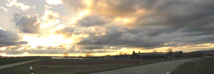 Das Bild zeigt eine Straße, bewölkten Himmel und die Sonne, die durch die Wolken bricht